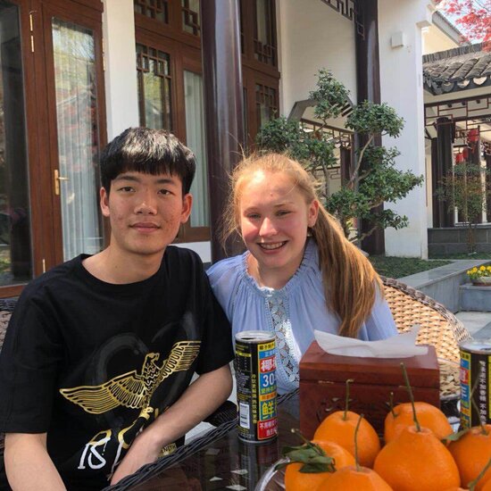 Eine deutsche Schülerin und ein chinesischer Schüler sitzen im Freien an einem Tisch vor einer Schale mit Orangen