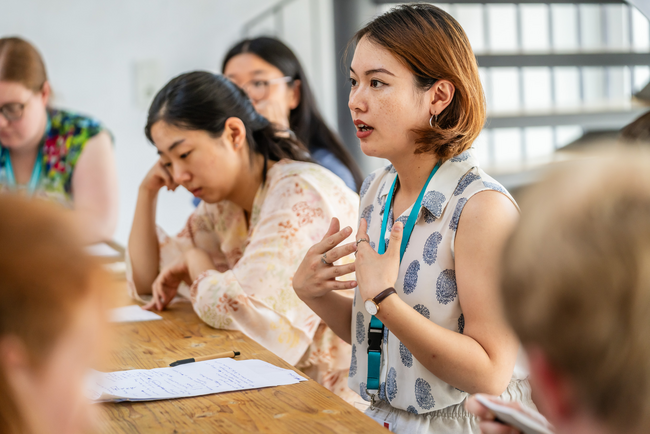 Mehrere asiatisch-gelesene und weiße Personen sitzen um einen Tisch herum. Eine Person spricht, die anderen hören zu